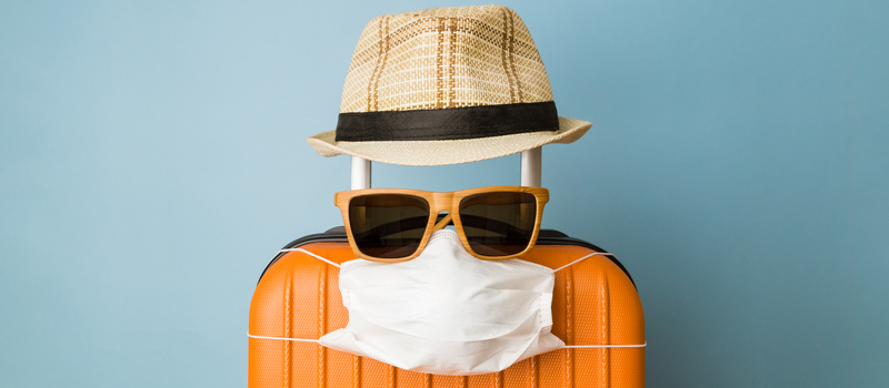 Suitcase with hat, sunglasses and protective medical mask on pastel blue background minimal creative coronavirus covid-19 travel concept.