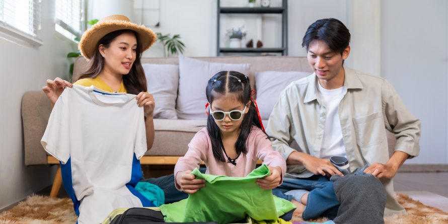 A girl packing her own clothes with her parents at the back, as part of their travel essentials list